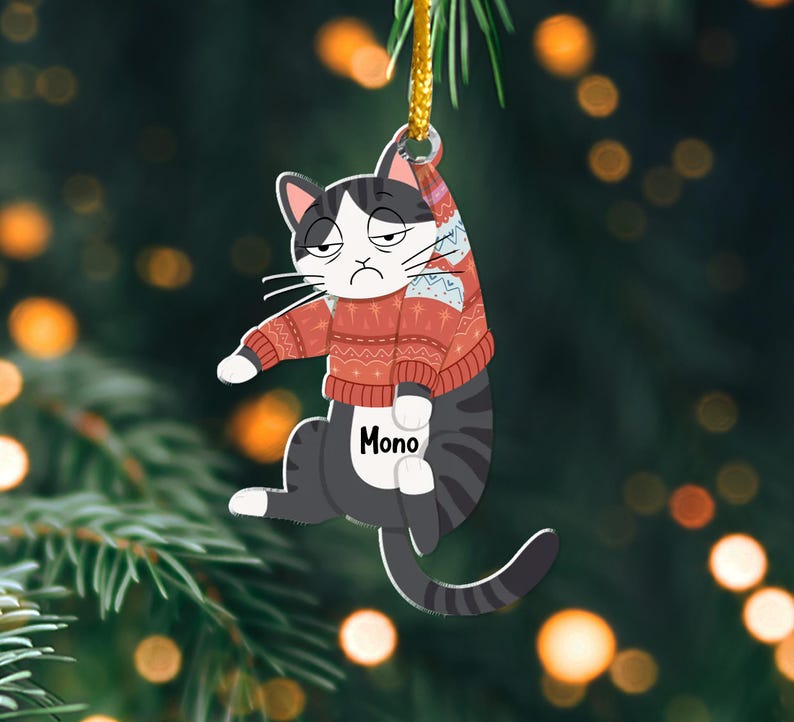 以下が含まれることがあります： 「Mono」と印刷された赤と白のセーターを着た白黒の猫。猫は金色の紐にぶら下がっていて、クリスマスのオーナメントです。