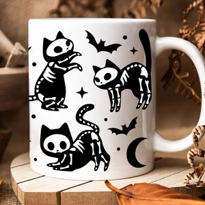 Pu&ograve; includere: Tazza in ceramica bianca con un'illustrazione in bianco e nero di tre gatti scheletro con lune crescenti e pipistrelli.