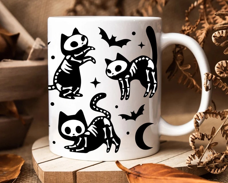 Pu&ograve; includere: Tazza in ceramica bianca con un'illustrazione in bianco e nero di tre gatti scheletro con lune crescenti e pipistrelli.