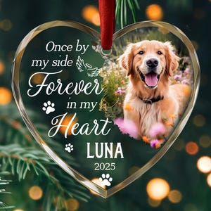 Puede incluir: Adorno de vidrio en forma de corazón con una foto de un perro golden retriever. El adorno tiene el texto "Once by my side, Forever in my Heart, Luna 2025". Se adjunta una cinta roja para colgar. El fondo está borroso.