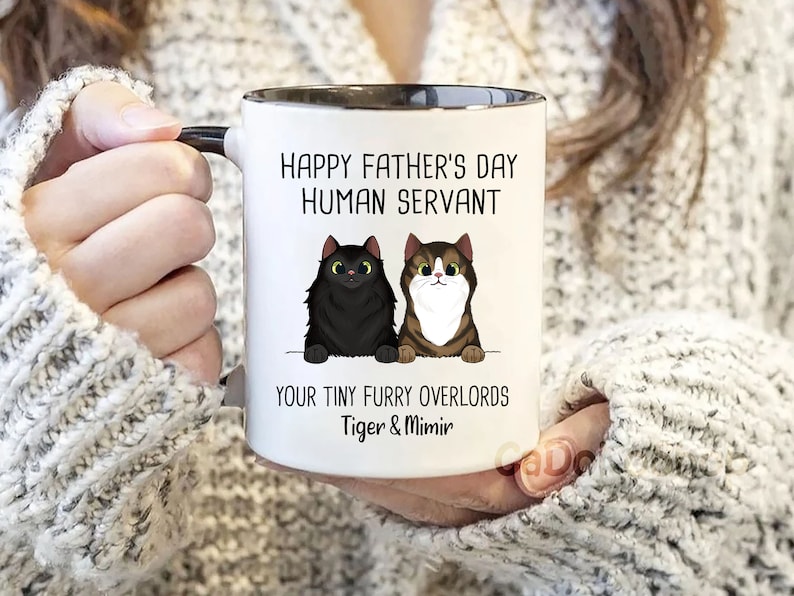 May include: White ceramic mug with a black rim and handle. The mug has a black and brown cat illustration and the text "Happy Father's Day Human Servant Your Tiny Furry Overlords Tiger & Mimir".