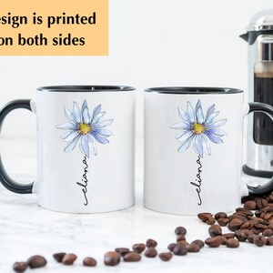 Puede incluir: Dos tazas de cer&aacute;mica blanca con asas negras. Cada taza presenta una margarita azul en acuarela con un centro amarillo y el nombre "Eliana" escrito verticalmente debajo de la flor. Las tazas est&aacute;n sobre una superficie blanca con granos de caf&eacute; esparcidos a su alrededor.