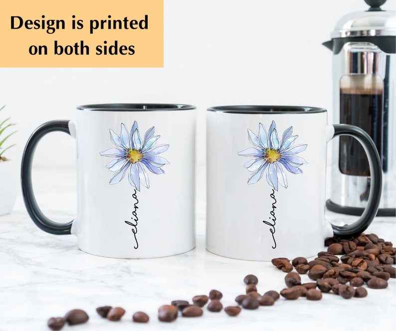 Puede incluir: Dos tazas de cer&aacute;mica blanca con asas negras. Cada taza presenta una margarita azul en acuarela con un centro amarillo y el nombre "Eliana" escrito verticalmente debajo de la flor. Las tazas est&aacute;n sobre una superficie blanca con granos de caf&eacute; esparcidos a su alrededor.
