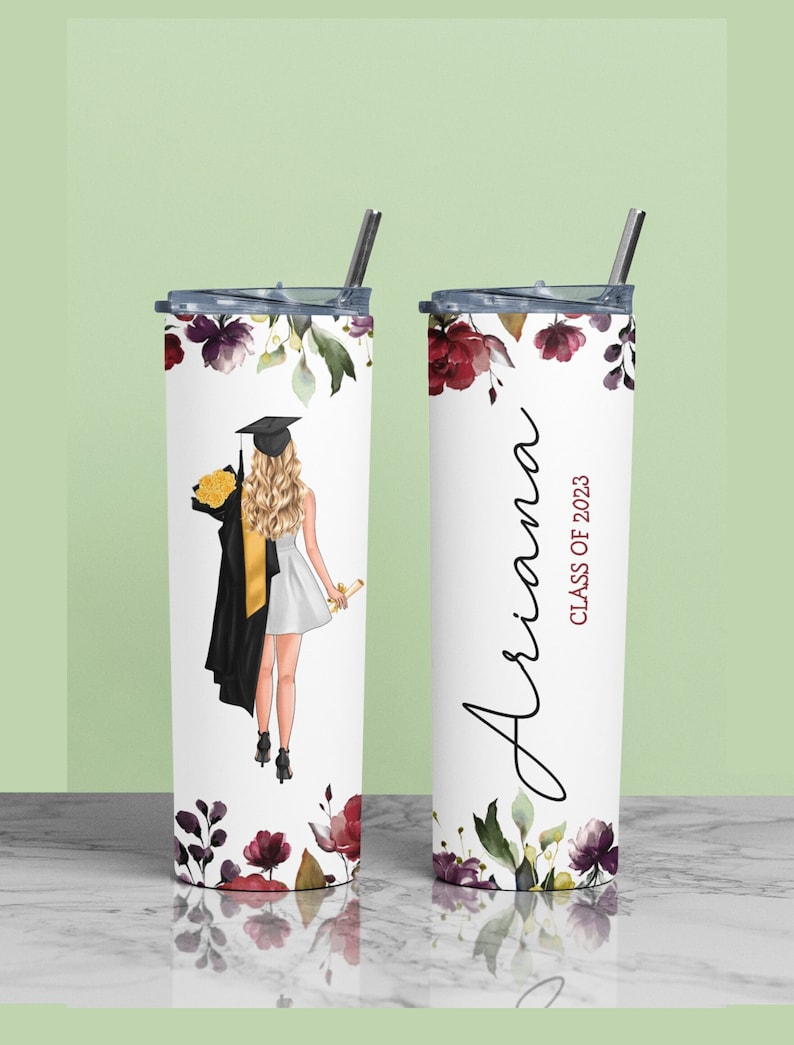 May include: A white stainless steel tumbler with a floral design and a graduation cap illustration. The tumbler has a black lid and a straw. The text "Ariana Class of 2023" is printed on the tumbler.