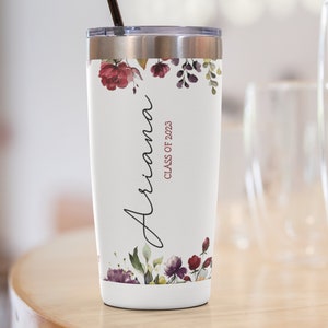 May include: A white stainless steel tumbler with a floral design and the name "Ariana" and the text "Class of 2023" in maroon. The tumbler has a black straw.