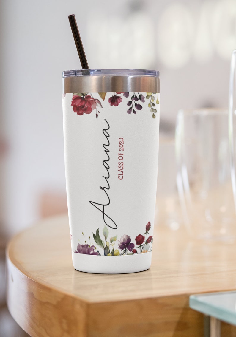 May include: A white stainless steel tumbler with a floral design and the name "Ariana" and the text "Class of 2023" in maroon. The tumbler has a black straw.