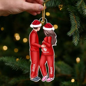 Op de afbeelding: Een kerstversiering met een stel in rode pyjama's en kerstmutsen. De vrouw is zwanger, met de tekst "Baby 2026" en voetafdrukken op haar buik. De namen "Robert" en "Ashley" staan op de pyjama's.