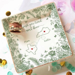 May include: Square ceramic trinket tray with a floral design in green and a gold rim. The tray features the text "wish you lived next door" and a map with two states and hearts. A rose gold ring sits on the tray.