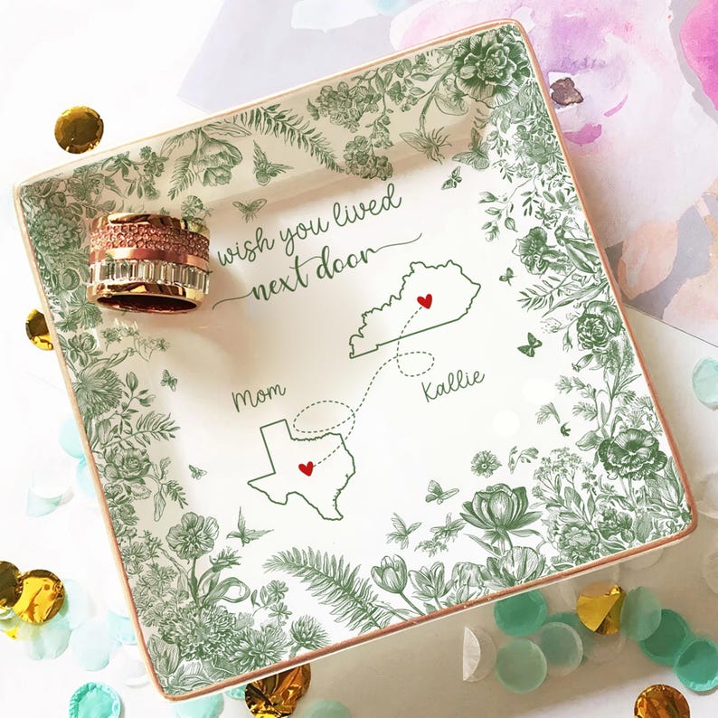 May include: Square ceramic trinket tray with a floral design in green and a gold rim. The tray features the text "wish you lived next door" and a map with two states and hearts. A rose gold ring sits on the tray.