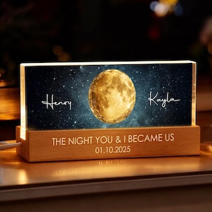 May include: A personalized night light with a wooden base and a rectangular illuminated panel. The panel features a golden moon, a starry background, and the names "Henry" and "Kayla". The text "THE NIGHT YOU & I BECAME US 01.10.2025" is also displayed.