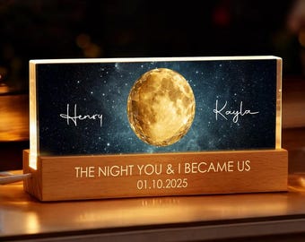 May include: A personalized night light with a wooden base and a rectangular illuminated panel. The panel features a golden moon, a starry background, and the names "Henry" and "Kayla". The text "THE NIGHT YOU & I BECAME US 01.10.2025" is also displayed.