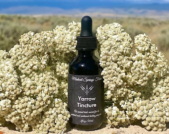 May include: A dark glass bottle with a black dropper top, labeled "Yarrow Tincture" with a black and white graphic design. The bottle is surrounded by white yarrow flowers and a blue sky with white clouds in the background.