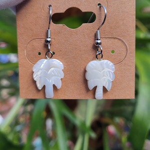 Peut inclure: Une paire de boucles d'oreilles pendantes argentées avec des breloques en forme de palmier blanc. Les palmiers sont faits d'un matériau blanc iridescent.