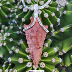 Può includere: Un ciondolo a forma di punta di freccia rosso e marrone avvolto in filo d'argento, appeso a una catena d'argento. Il ciondolo è mostrato su uno sfondo di cactus verde.