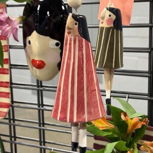 May include: Ceramic art pieces suspended on a metal grid. The items include a black face vase with red lips, and two doll-like figures with striped dresses in red and green. The figures have black hair and painted faces.