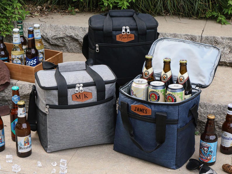 Puede incluir: Tres bolsas isot&eacute;rmicas personalizadas con cierre de cremallera. Las bolsas son grises, negras y azules. Las bolsas tienen parches de cuero con las iniciales "MJK", "Martin" y "James" en ellas. Las bolsas est&aacute;n llenas de botellas de cerveza.