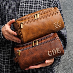 Könnte beinhalten: Zwei braune Kulturbeutel aus Leder mit goldenen Reißverschlüssen und personalisierten Initialen. Auf der oberen Tasche steht "KENNETH", auf der unteren Tasche "CDE". Die Taschen werden von einer Person gehalten, die ein kariertes Hemd trägt.