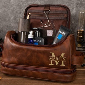 Könnte beinhalten: Eine braune Leder-Kulturtasche mit Doppelreißverschluss, gefüllt mit Pflegeprodukten. Die Tasche ist mit dem Namen "MARTIN" und der römischen Zahl "IV" geprägt. Im Inneren befinden sich Flaschen, eine Tube "Man'SHK" Creme und ein Holzbehälter.