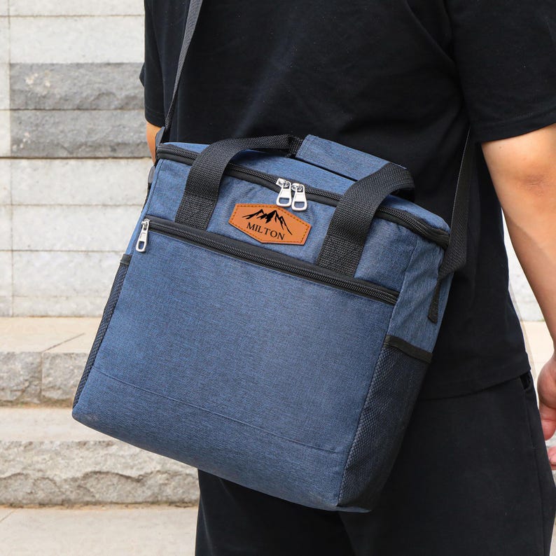 Puede incluir: Una bolsa de almuerzo azul aislada con una cremallera negra y un parche de cuero marr&oacute;n que dice "MILTON" con un dise&ntilde;o de cordillera. La bolsa tiene una correa para el hombro y dos asas superiores.