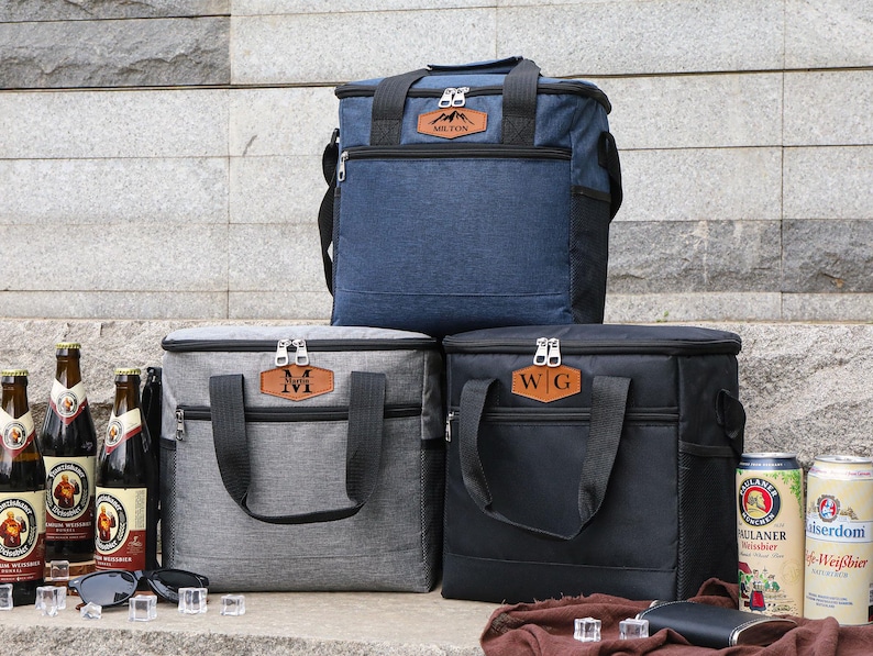 Puede incluir: Tres bolsas de almuerzo aisladas con iniciales personalizadas. Las bolsas son grises, negras y azules. Las bolsas est&aacute;n sobre una superficie de piedra con botellas de cerveza delante.