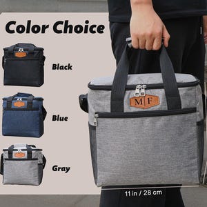 Puede incluir: Una bolsa de almuerzo aislada gris con un parche de cuero marr&oacute;n que dice "MF". La bolsa tiene un cierre de cremallera y un asa negra. La bolsa mide 28 cm de ancho y 16 cm de alto. La bolsa est&aacute; disponible en negro, azul y gris.