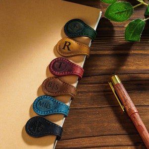 May include: A notebook with six leather bookmarks. The bookmarks are different colors and have initials on them. The initials are "R", "T", "B|S", "MNS", and "MH". A wooden pen with a gold tip is on the table next to the notebook.