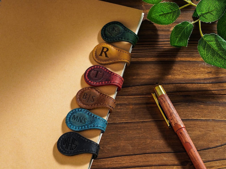 May include: A notebook with six leather bookmarks. The bookmarks are different colors and have initials on them. The initials are "R", "T", "B|S", "MNS", and "MH". A wooden pen with a gold tip is on the table next to the notebook.