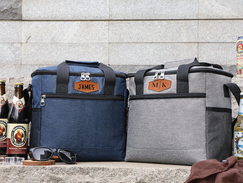 Puede incluir: Dos bolsas de almuerzo aisladas personalizadas, una azul y otra gris, con etiquetas de nombre de cuero. La bolsa azul tiene el nombre "JAMES" y la bolsa gris tiene las iniciales "M/K".