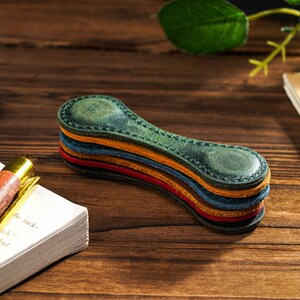 May include: A stack of four leather bookmark magnets in shades of green, orange, blue, and red. The magnets are shaped like a long, thin oval and have a subtle textured finish.