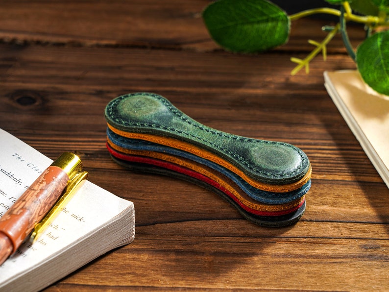 May include: A stack of four leather bookmark magnets in shades of green, orange, blue, and red. The magnets are shaped like a long, thin oval and have a subtle textured finish.