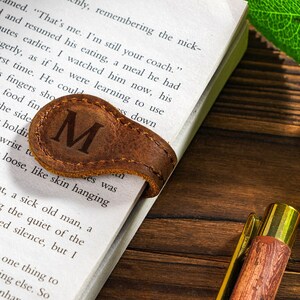 May include: A brown leather bookmark with the letter "M" embossed on it. The bookmark is placed on a book with text visible. The book is on a wooden surface.