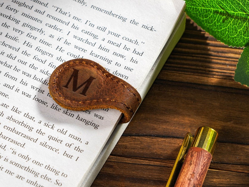 May include: A brown leather bookmark with the letter "M" embossed on it. The bookmark is placed on a book with text visible. The book is on a wooden surface.