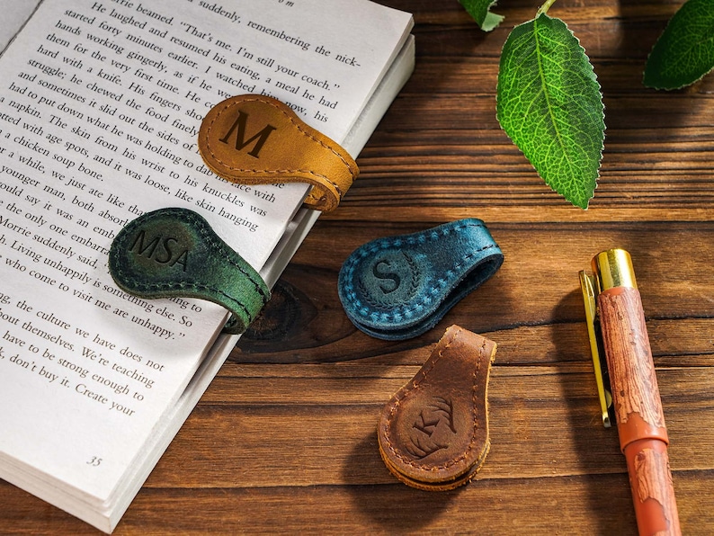 May include: Three leather book markers with personalized initials. The markers are in brown, green, and blue leather. The initials are "M", "MSA", and "S".