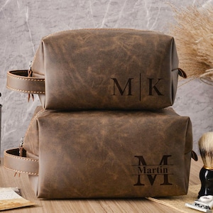 Puede incluir: Dos neceseres de cuero marrón apilados. La bolsa superior tiene las iniciales "MK" grabadas en la parte delantera. La bolsa inferior tiene el nombre "Martin" y los números romanos "IVI" grabados.