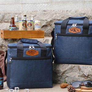Puede incluir: Dos bolsas isot&eacute;rmicas de lona azul con etiquetas de nombre personalizadas de cuero marr&oacute;n. La bolsa m&aacute;s grande tiene las iniciales "MKS" y la bolsa m&aacute;s peque&ntilde;a tiene el nombre "MARTIN". Ambas bolsas est&aacute;n sobre una superficie de piedra con algunas botellas de cerveza en el fondo.
