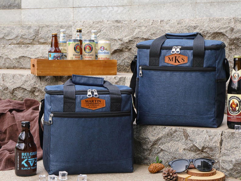 Puede incluir: Dos bolsas isot&eacute;rmicas de lona azul con etiquetas de nombre personalizadas de cuero marr&oacute;n. La bolsa m&aacute;s grande tiene las iniciales "MKS" y la bolsa m&aacute;s peque&ntilde;a tiene el nombre "MARTIN". Ambas bolsas est&aacute;n sobre una superficie de piedra con algunas botellas de cerveza en el fondo.