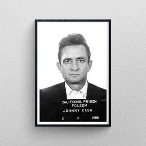 May include: Black and white mugshot of Johnny Cash, taken at California Prison Folsom in 1966. The mugshot shows Cash with a serious expression, wearing a suit jacket and tie. The text "CALIFORNIA PRISON FOLSOM JOHNNY CASH 11 8 1966" is printed below the image.