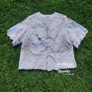 May include: A white short-sleeved blouse featuring a large scalloped collar, brown floral embroidery, and three front tie closures. The garment includes a label reading "jen byrne" at the neckline and a watermark logo "jen byrne studio nine".
