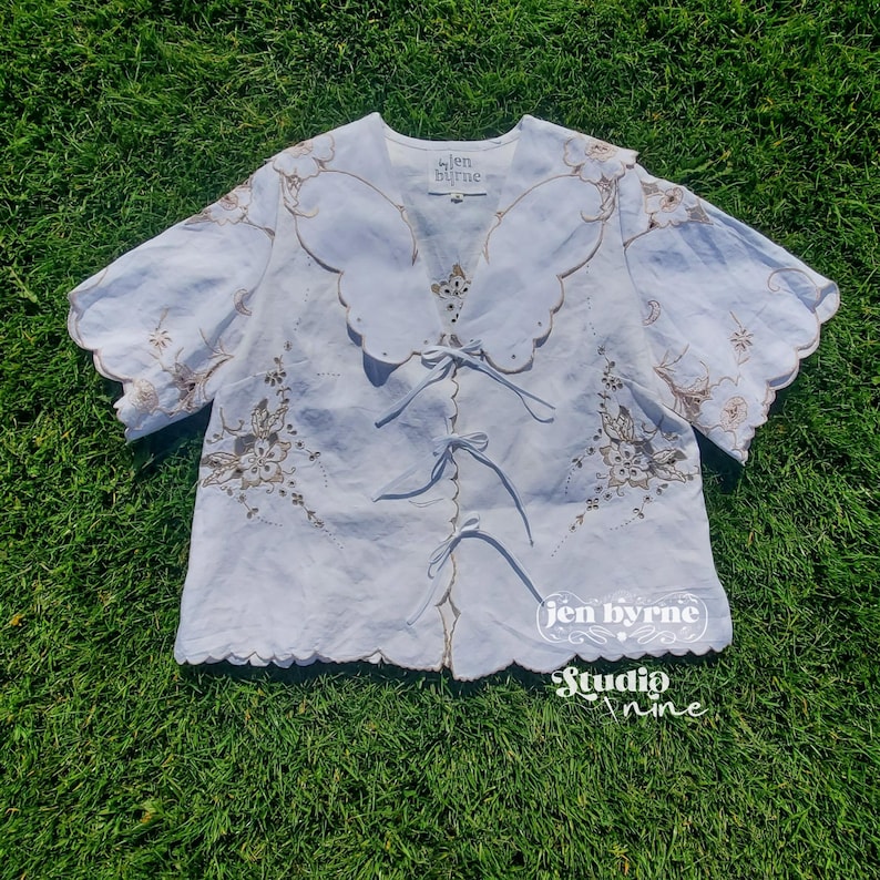 May include: A white short-sleeved blouse featuring a large scalloped collar, brown floral embroidery, and three front tie closures. The garment includes a label reading "jen byrne" at the neckline and a watermark logo "jen byrne studio nine".