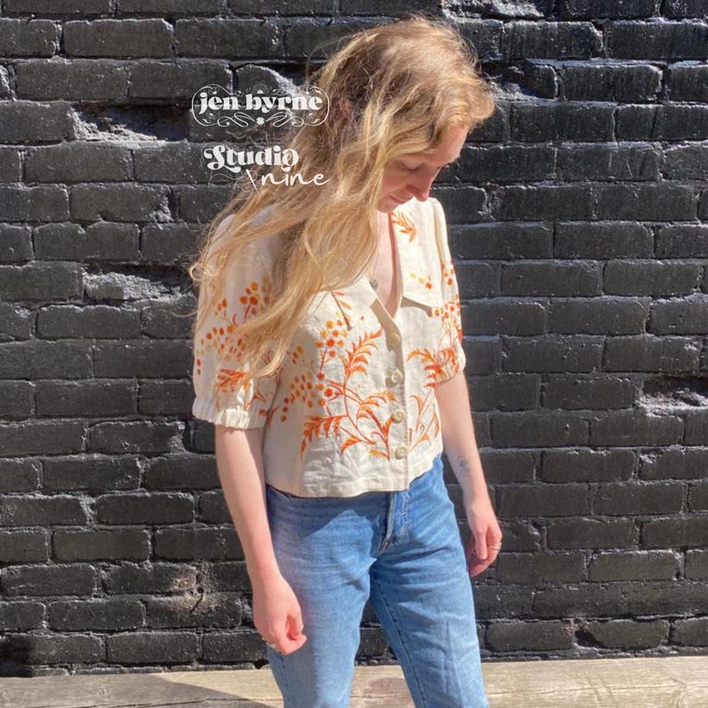 May include: A cream-colored short-sleeve button-down shirt featuring an orange floral and leaf pattern. The shirt has a collar and a front button closure. The text "jen byrne Studio nine" is visible in the upper left corner of the image.