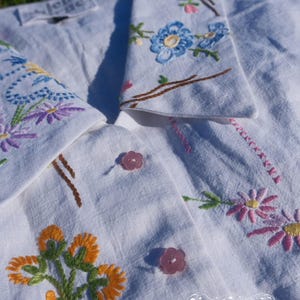 May include: A white button-down shirt featuring colorful floral embroidery on the collar and front. The shirt has light pink flower-shaped buttons and includes the text "jen byrne Studio nine" printed in white on the lower right side.