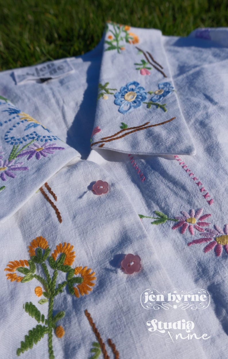 May include: A white button-down shirt featuring colorful floral embroidery on the collar and front. The shirt has light pink flower-shaped buttons and includes the text "jen byrne Studio nine" printed in white on the lower right side.