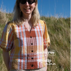 May include: A short-sleeved button-down blouse featuring a plaid pattern in shades of yellow, orange, and white with a textured fabric finish. The blouse has a V-neckline and dark buttons. Text on the image reads jen byrne Studio nine.