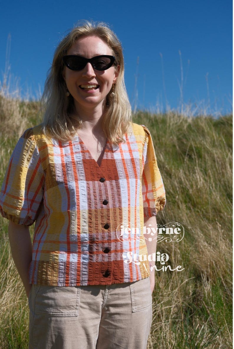 May include: A short-sleeved button-down blouse featuring a plaid pattern in shades of yellow, orange, and white with a textured fabric finish. The blouse has a V-neckline and dark buttons. Text on the image reads jen byrne Studio nine.