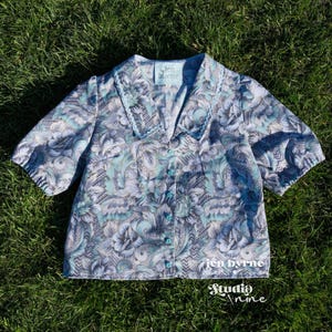 May include: A short-sleeved button-down blouse featuring a light blue, grey, and white floral paisley pattern with a wide collar and light blue trim. The inner label reads "jen byrne" and the bottom right corner displays the text "jen byrne Studio nine".