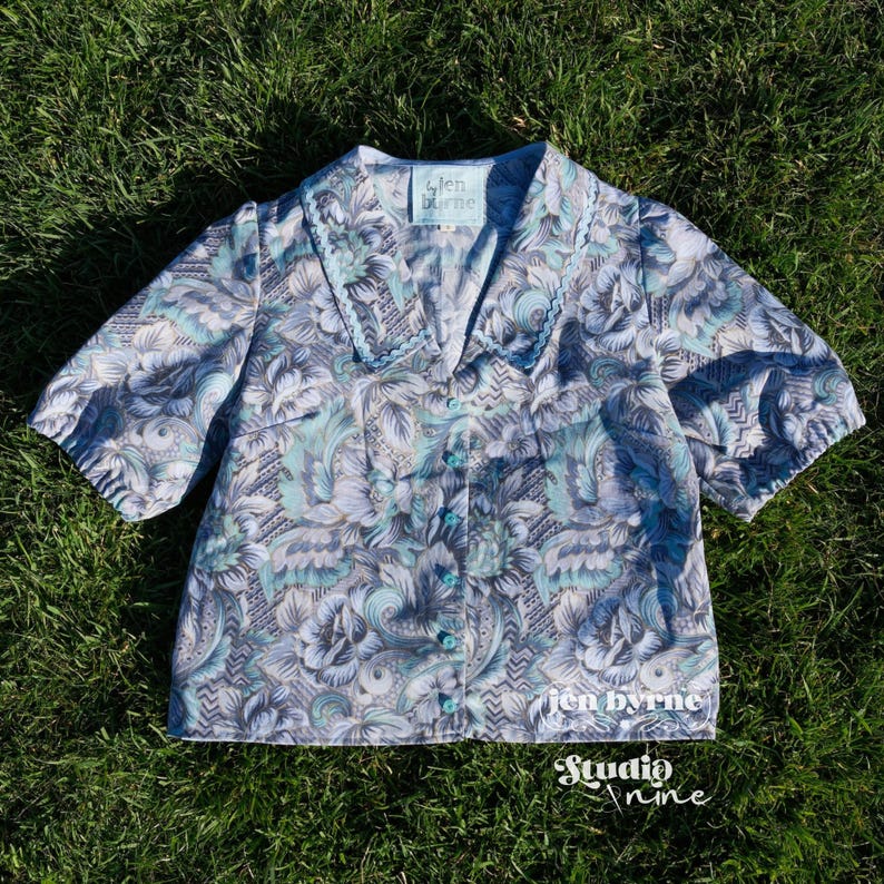 May include: A short-sleeved button-down blouse featuring a light blue, grey, and white floral paisley pattern with a wide collar and light blue trim. The inner label reads "jen byrne" and the bottom right corner displays the text "jen byrne Studio nine".