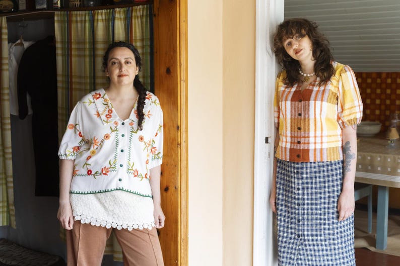 May include: A white blouse with floral embroidery and a button-down front, paired with brown trousers. Beside it, a yellow and orange plaid short-sleeved blouse with a button-down front, worn with a blue and white plaid button-down midi skirt.