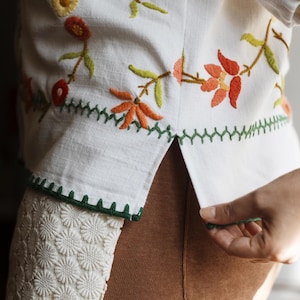 May include: A white shirt featuring colorful floral embroidery in shades of orange, yellow, and green. The hem includes a green zigzag stitch detail and a white floral lace trim along the side. The shirt is worn over brown pants.