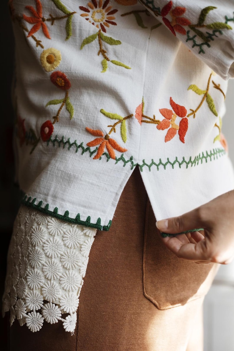 May include: A white shirt featuring colorful floral embroidery in shades of orange, yellow, and green. The hem includes a green zigzag stitch detail and a white floral lace trim along the side. The shirt is worn over brown pants.