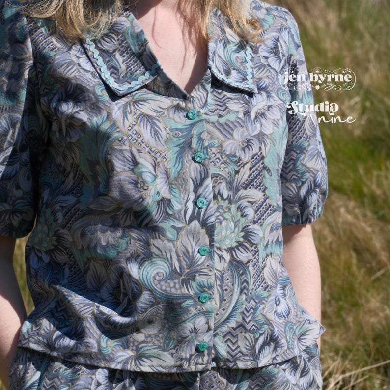 May include: A short-sleeved button-down shirt featuring a blue, gray, and teal floral pattern with a notched collar. The shirt has a row of teal buttons down the front and includes the text "jen byrne" and "studio nine" on the right side.